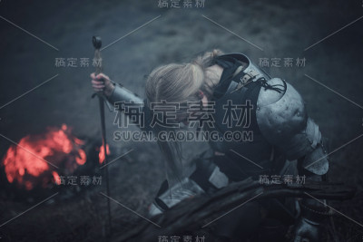 《贞德》中的女孩身披铠甲，双手握剑，跪在火与烟的背景下。