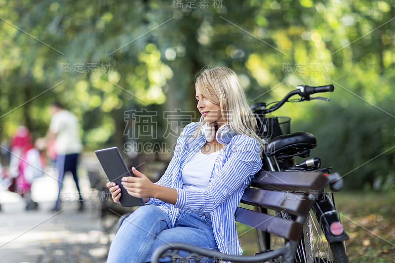 金发女人坐在长椅上，手上拿着平板电脑，脖子上戴着耳机