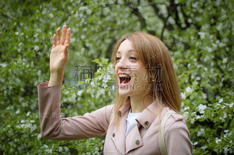 年轻美丽的女人与金发说你好某人在一个大开花树在一个春天公园，户外女性肖像