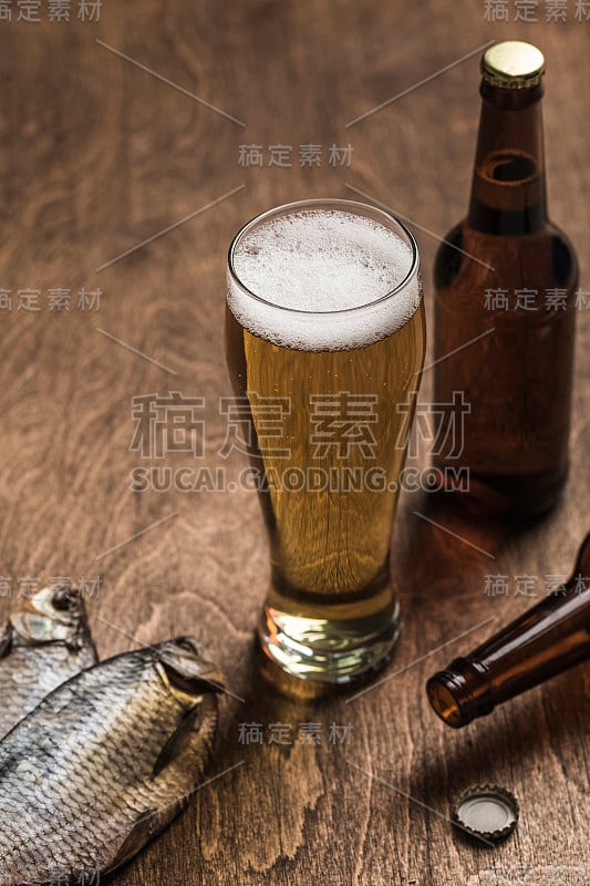 两个深色啤酒瓶，一杯啤酒和泡沫，木色背景上放着干鱼。特写镜头。