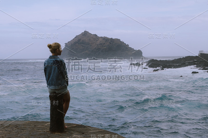 年轻女子坐在岩石上，看着汹涌的大海和破浪