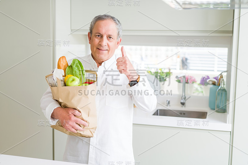 英俊的老男人拿着一个纸袋新鲜的食品杂货在厨房快乐与大微笑做ok的手势，拇指与手指向上，优秀的手势