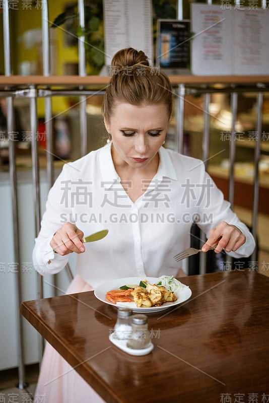 一位身穿白衬衫的妇女正坐在餐桌旁用餐，手里拿着刀叉