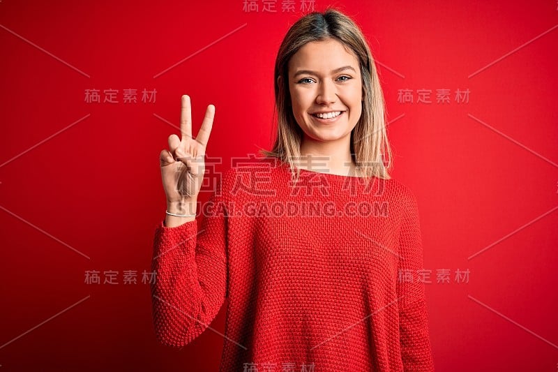 年轻美丽的金发女人穿着休闲毛衣在红色孤立的背景显示和指向第2个手指，而微笑自信和快乐。