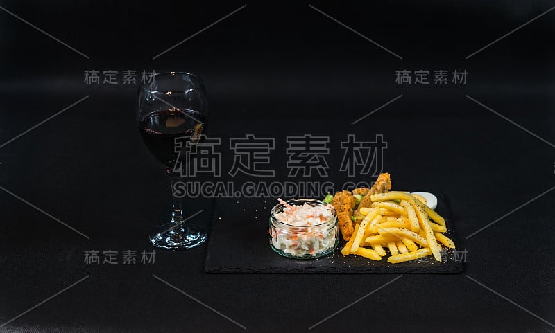 这道菜由裹着面包屑的鸡胸肉、炸薯条和沙拉组成，杯中红酒，黑色石制盘子