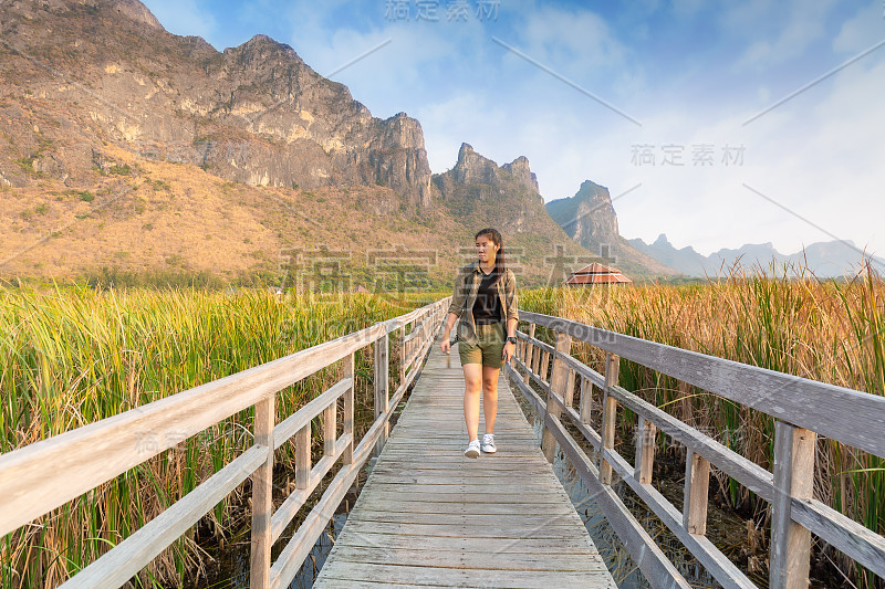 亚洲徒步旅行者背着沉重的背包，在一个小亭子户外徒步小径上的木桥上，沼泽与草地和蓝山背景。Khao S