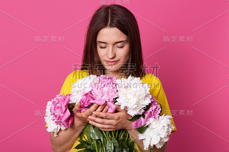 室内拍摄的美丽的黑发女性抱着大花束的粉红色和白色的牡丹花，有温柔的外观，在粉红色的工作室背景中摆出孤