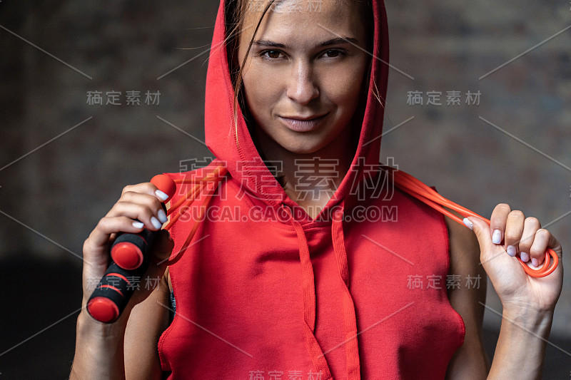 特写肖像漂亮的微笑运动的年轻女人在红色帽衫运动装抱跳绳跳绳绕脖子快乐看镜头砖墙背景减肥心脏健康的生活