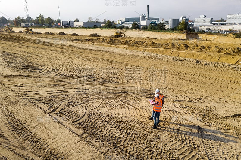 图为两名身穿橙色背心、戴着防护头盔的筑路工人在场地中间的俯视图