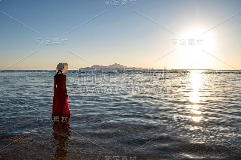 一名年轻女子身穿红色长裙，头戴草帽，站在海边欣赏初夏的朝阳。