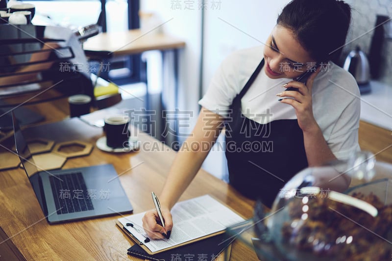 年轻可爱的学生在课后在咖啡店做服务员。白人时髦女孩与朋友打电话和写电话号码在餐厅新送美味蛋糕