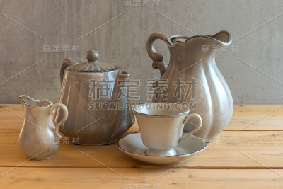 茶具是灰色的，放在木制的背景上。茶壶，奶精，杯子和茶碟在桌子上。陶瓷厨具