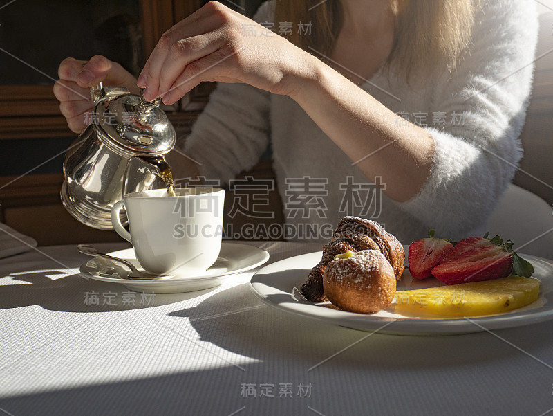 女孩在早餐时把茶倒进杯子里，盘子里有水果