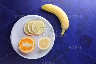极简主义风格的紫外光背景柑橘，白色盘子上的柠檬片和半片橙子，深蓝色背景上的黄色香蕉，健康食品，素食，