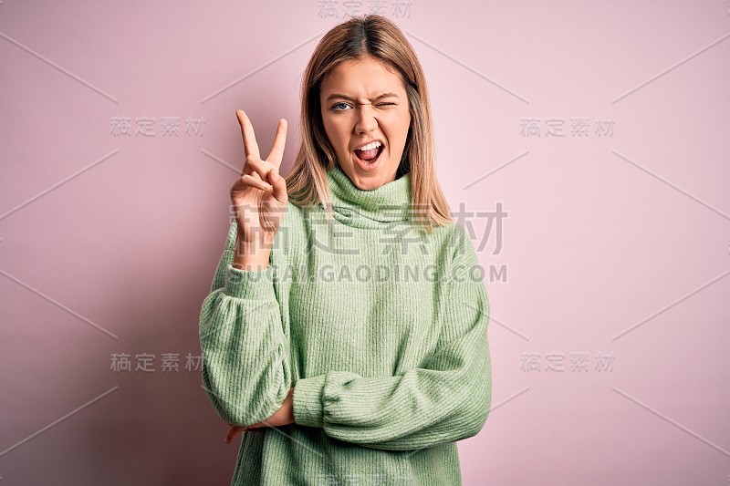 年轻美丽的金发女人穿着冬季羊毛衫在粉红色孤立的背景微笑与快乐的脸在镜头做胜利标志。二号人物。