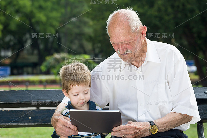 夏天，爷爷和孙子在公园的长凳上玩平板电脑