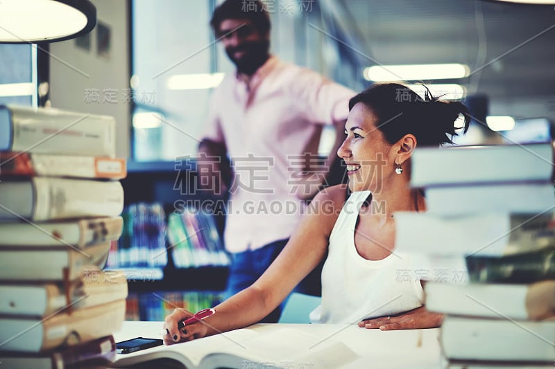 微笑的大学生坐在书桌与研究书籍在大学图书馆，年轻的亚洲女性有乐趣与她的男同学在现代高级商学院的学习大