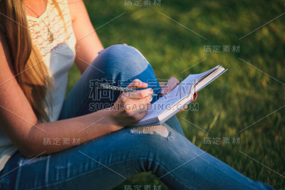 女孩文案坐在草地上，拿着笔记本和笔。在户外写作的女性自由职业者。