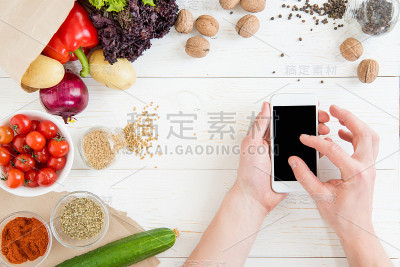 近距离观看的人使用智能手机与空白屏幕，而烹饪和新鲜的原料在木桌上