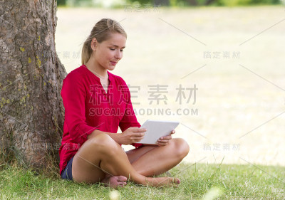 快乐的年轻女子坐在草地上研究无线平板电脑