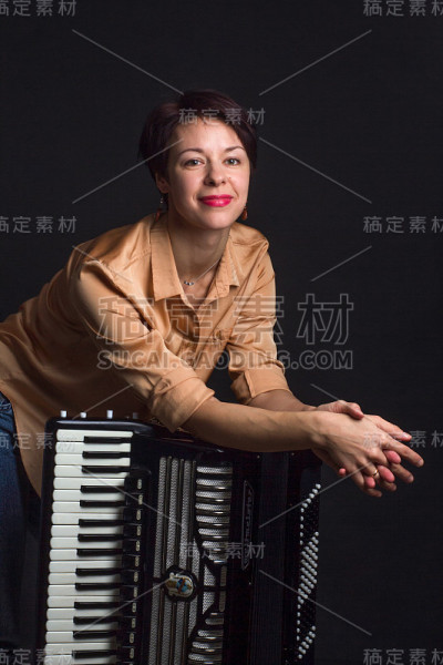 一个穿着金色衬衫、留着短发的黑发女人站在工作室的黑暗背景上。与手风琴的音乐家