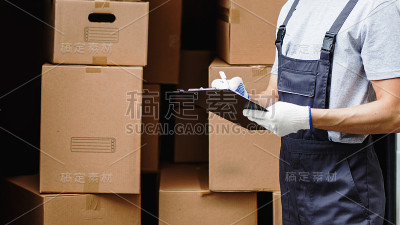 一位穿着制服、面带微笑的年轻工人站在装满箱子的货车旁，手里拿着一块剪贴板。搬家，搬家服务