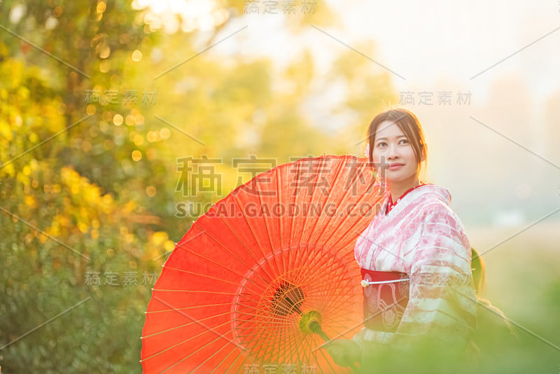 快乐年轻迷人美丽的亚洲女人的肖像女孩穿着粉红色的传统日本和服拿红纸伞微笑与树叶在秋天在东京城市，日本