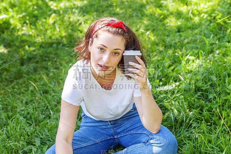 快乐的女孩微笑着在户外吃午饭。美丽年轻的黑发女子与带走的咖啡杯休息在公园或花园绿地的背景。教育自由职