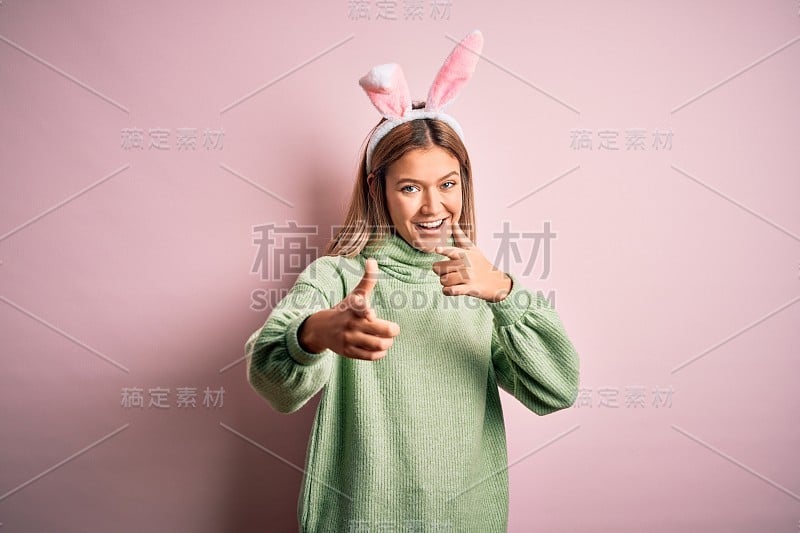 年轻美丽的女人戴着复活节兔子耳朵站在孤立的粉红色背景指向手指与快乐和有趣的脸相机。良好的能量和共鸣。