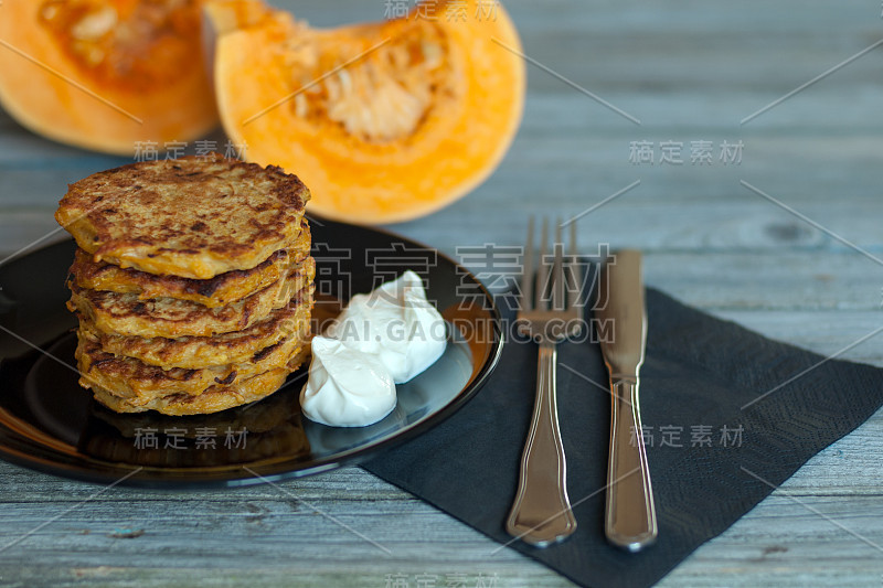 黑色的盘子上放着天然的酸奶酪煎饼，灰色的木桌上放着刀叉餐巾，背景是橘色的南瓜