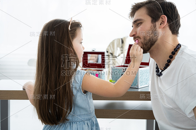 可爱的小女儿和戴着皇冠的帅气爸爸在孩子的房间里一起玩耍。女孩在给她爸爸化妆。