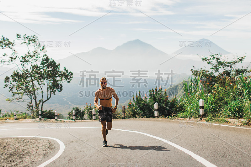 在炎热的夏天，男人在上坡的柏油路上赤裸上身跑步。全景山景背景。使用胸心率监测器。