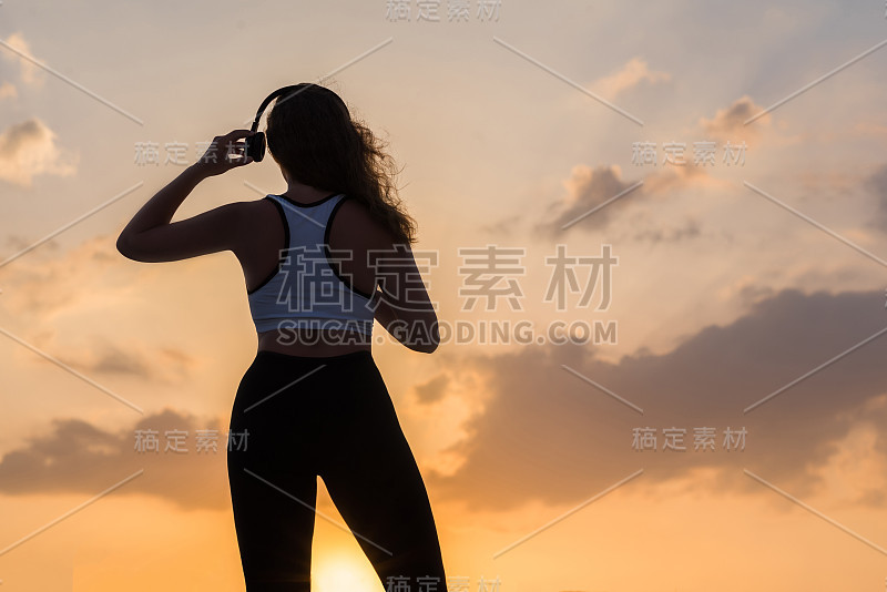 年轻女子的剪影穿着运动服，耳机上听着夕阳天空下的音乐背景。健康生活方式的概念