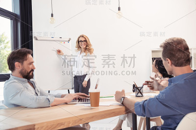 带着眼镜微笑的女商人指着空白的白板，看着同事
