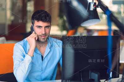 年轻的商人在办公室一边忙着用笔记本电脑一边用智能手机聊天。