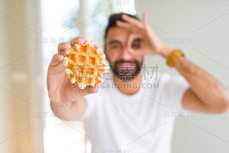 英俊的西班牙男子吃甜蜜的比利时华夫饼与快乐的脸微笑做ok手势与手在眼睛透过手指