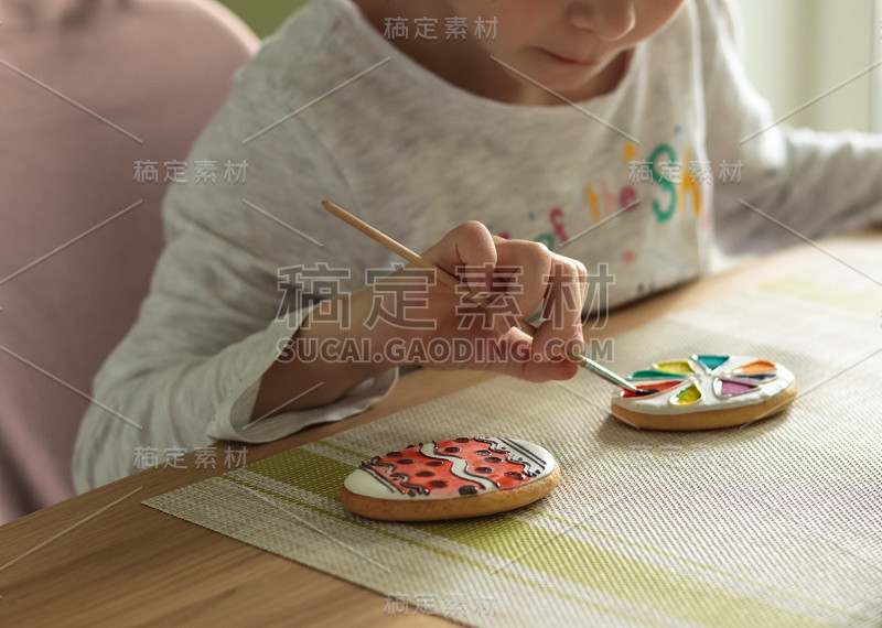 一个扎着辫子的小女孩坐在厨房的桌前，用画笔在复活节饼干上涂颜料